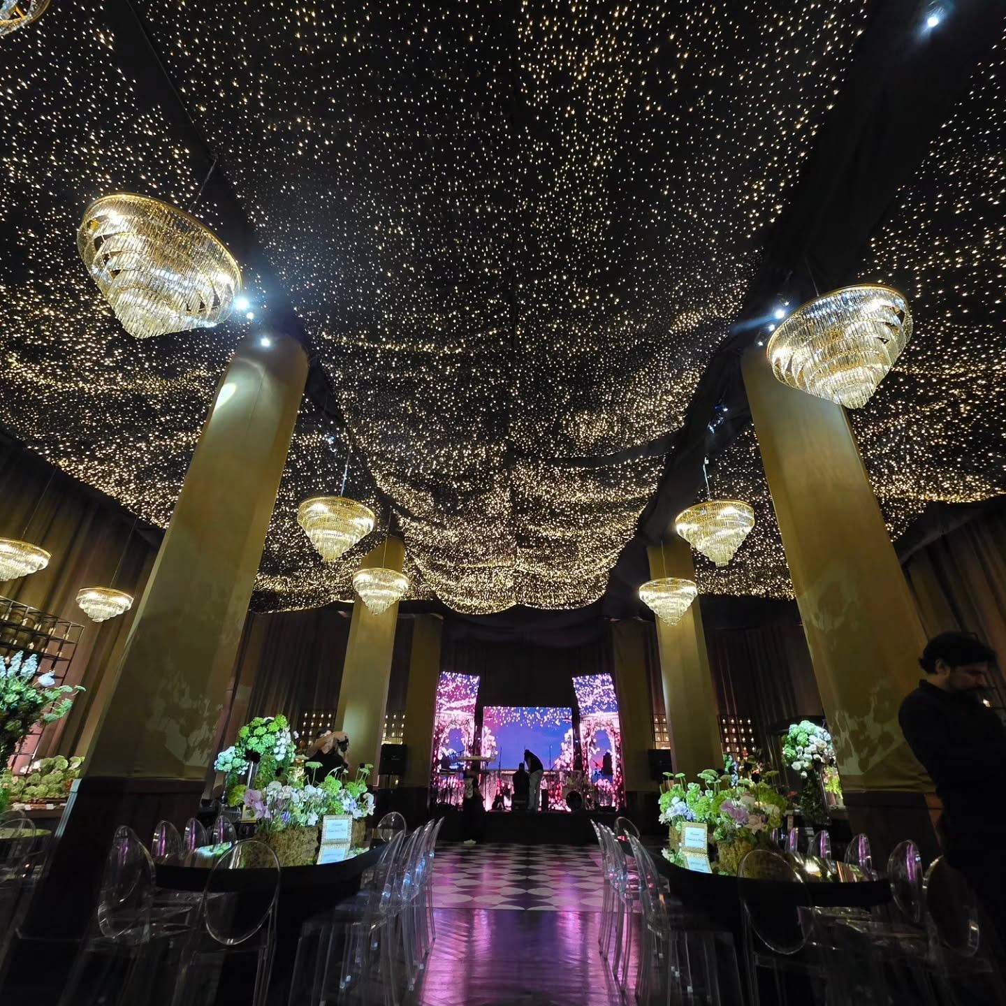 Salón de evento con techo de luces, candelabros de cristal y escenario con pantallas al fondo